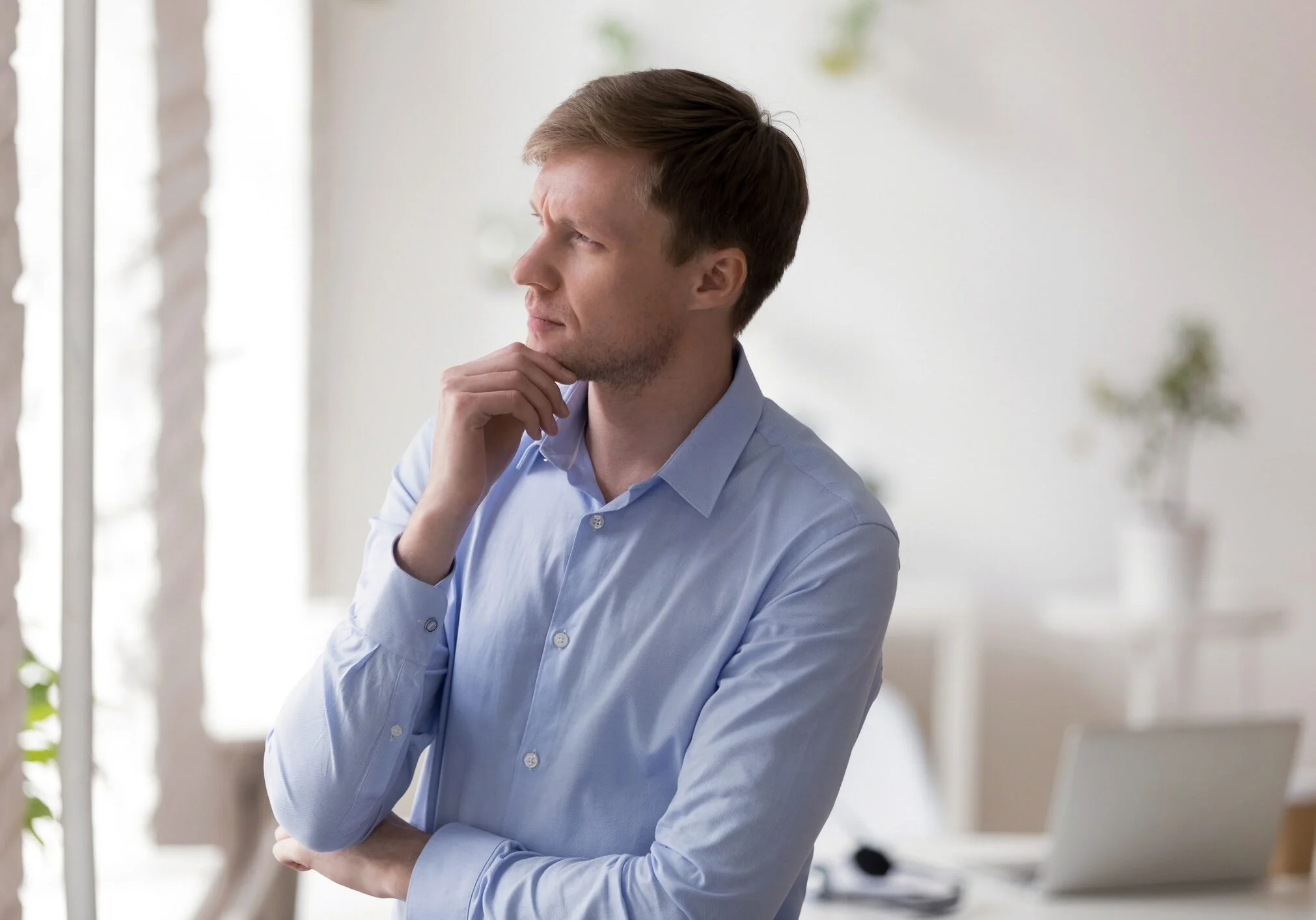 Serious concentrated millennial businessman standing alone in modern office room looking away touching stroking chin thinking about problem or making important decision planning new future projects